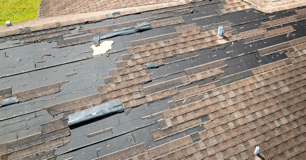 Weathered residential roof in MD with significant damage. Large sections of the brown asphalt shingles are missing, exposing the underlying dark roofing felt. Displaced metal flashing and visible roof vents indicate deterioration and the need for repair.
