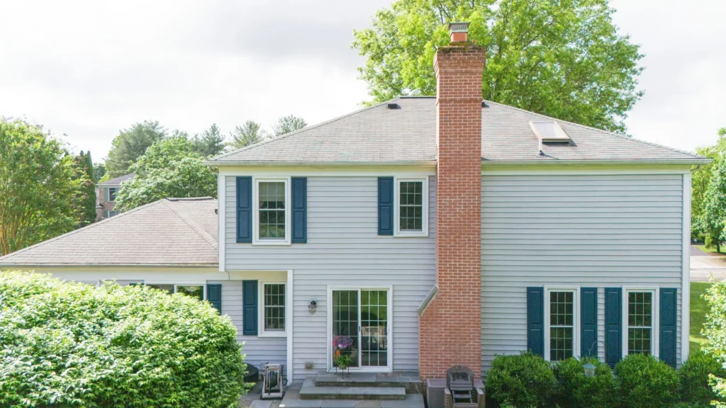 Grey asphalt shingle roof and blue shuttered windows newly installed to a red-brick house surrounded by lush green lawn and tress in the suburbs of Maryland.